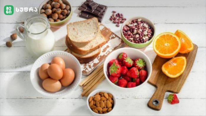 Assorted breakfast ingredients on a white wooden table: eggs, milk, sliced bread, almonds, chocolate, pistachios, strawberries, oats, and orange halves.