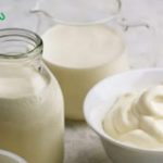 Milk bottles and a pitcher of milk beside a bowl of whipped cream on a light countertop, with a brand logo in the top-left corner.