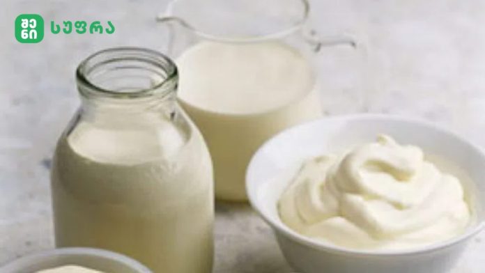 Milk bottles and a pitcher of milk beside a bowl of whipped cream on a light countertop, with a brand logo in the top-left corner.