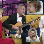 A man in a tuxedo plays an acoustic guitar on a colorful TV talk-show set, flanked by two smiling women holding a baby on a yellow couch.