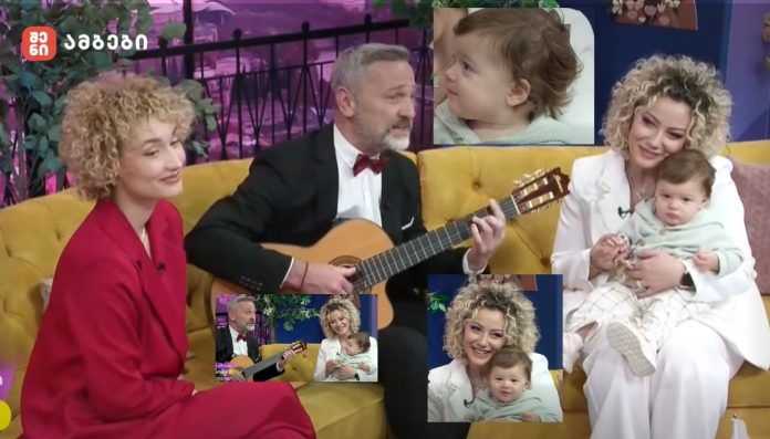 A man in a tuxedo plays an acoustic guitar on a colorful TV talk-show set, flanked by two smiling women holding a baby on a yellow couch.