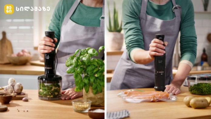 Two people in green shirts and gray aprons using a handheld blender in a bright kitchen; left side herbs and garlic on the counter, right side salmon being prepared with an immersion blender in front of them.