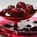 Clear glass bowl filled with bright red juice and dark fruit pieces, likely cherries, on a pale background.