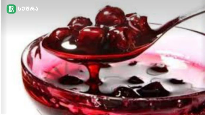 Clear glass bowl filled with bright red juice and dark fruit pieces, likely cherries, on a pale background.