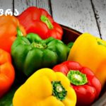 Assorted bell peppers (orange, green, red, yellow) in a bowl on a white wooden table, with a logo in the corner.