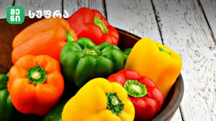 Assorted bell peppers (orange, green, red, yellow) in a bowl on a white wooden table, with a logo in the corner.