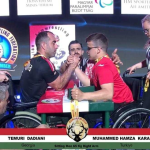 Two wheelchair arm wrestlers face off on a stage, gripping hands as referees observe; banners and flags of Georgia and Turkey appear in the background.