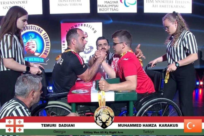 Two wheelchair arm wrestlers face off on a stage, gripping hands as referees observe; banners and flags of Georgia and Turkey appear in the background.