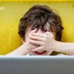 Child covers eyes with hands while looking at a laptop in front of a yellow wall.