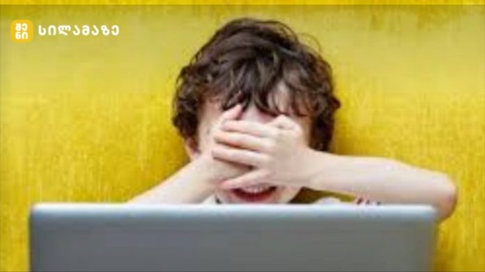 Child covers eyes with hands while looking at a laptop in front of a yellow wall.