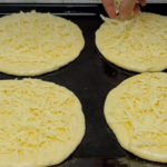 Four round dough discs topped with shredded cheese on a baking sheet; a hand is sprinkling cheese on the top left piece.