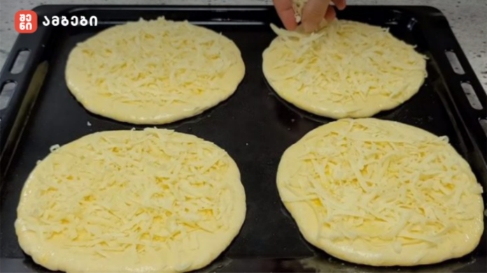 Four round dough discs topped with shredded cheese on a baking sheet; a hand is sprinkling cheese on the top left piece.