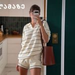 Person taking a mirror selfie in a modern white kitchen, wearing cream striped loungewear and carrying a red bag.
