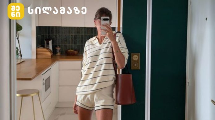 Person taking a mirror selfie in a modern white kitchen, wearing cream striped loungewear and carrying a red bag.