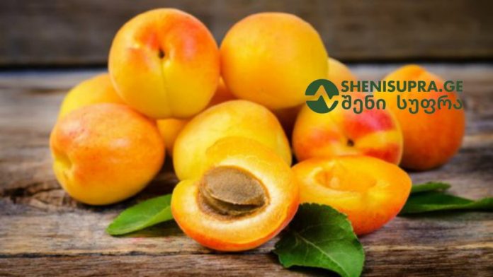 Pile of ripe apricots on a rustic wooden surface, with one cut open to show the stone and a couple of green leaves nearby in a natural setting.