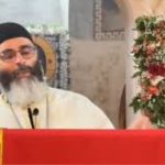 Orthodox priest with a black hat and glasses speaks at a red lectern, with Christ icon and floral garlands nearby.
