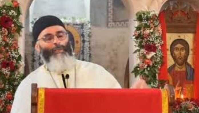 Orthodox priest with a black hat and glasses speaks at a red lectern, with Christ icon and floral garlands nearby.
