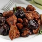 Plate of fried meat chunks mixed with dark prunes, garnished with a rosemary sprig and a cinnamon stick on the side.