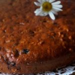 Close-up of a round golden-brown cake on a lace doily, adorned with white daisies.