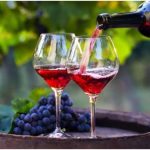 Two red wine glasses on a wooden barrel with wine being poured from a bottle, grapes and vines in the background.