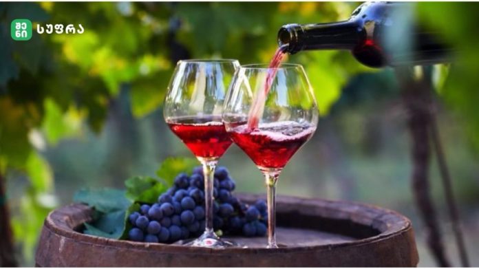 Two red wine glasses on a wooden barrel with wine being poured from a bottle, grapes and vines in the background.