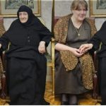 Two women sit indoors on chairs, one wearing a black hijab and dress and the other in a brown patterned shawl, holding hands.
