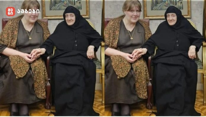 Two women sit indoors on chairs, one wearing a black hijab and dress and the other in a brown patterned shawl, holding hands.