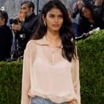 Woman with long dark hair walking the red carpet in a sheer beige blouse; photographers and a green hedge backdrop surround her.
