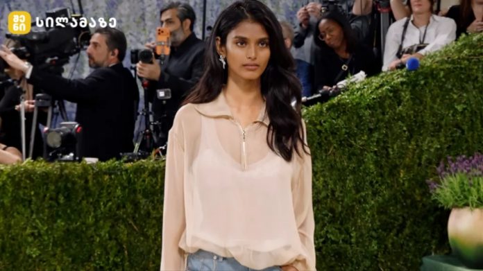 Woman with long dark hair walking the red carpet in a sheer beige blouse; photographers and a green hedge backdrop surround her.