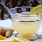 Glass mug of lemon tea with a lemon wedge, surrounded by ginger and orange slices on a white table in a snowy backdrop.