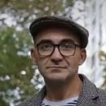 Portrait of a man wearing glasses and a flat cap outdoors, with a blurred fountain and greenery behind him; red logo in top-left corner.