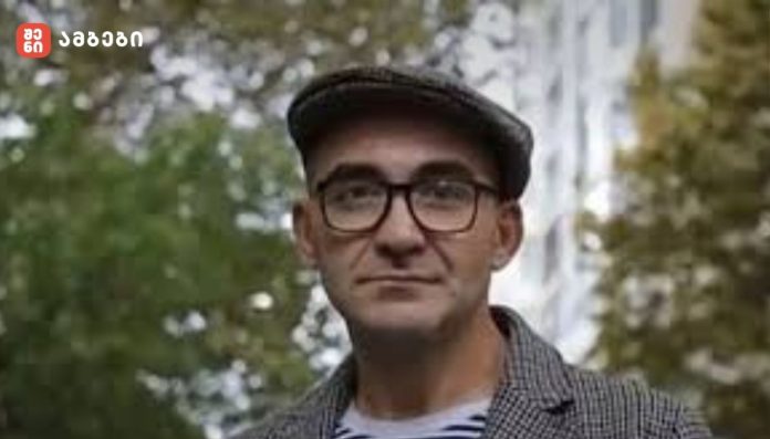 Portrait of a man wearing glasses and a flat cap outdoors, with a blurred fountain and greenery behind him; red logo in top-left corner.