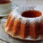 Bundt cake dusted with powdered sugar on a glass platter beside tea cups.