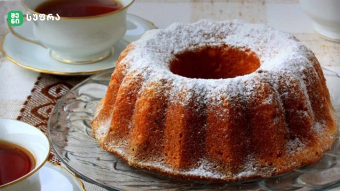 Bundt cake dusted with powdered sugar on a glass platter beside tea cups.
