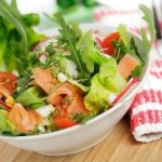 Bowl of mixed greens salad with tomatoes and smoked salmon, on a wooden table with a red-and-white striped napkin and fork nearby