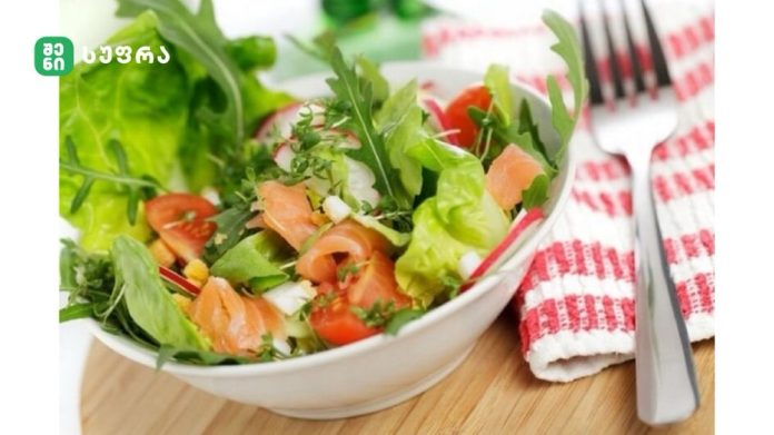Bowl of mixed greens salad with tomatoes and smoked salmon, on a wooden table with a red-and-white striped napkin and fork nearby