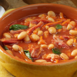 Yellow ceramic bowl holds a hearty white bean and tomato soup with greens on a wooden table, spoon nearby.