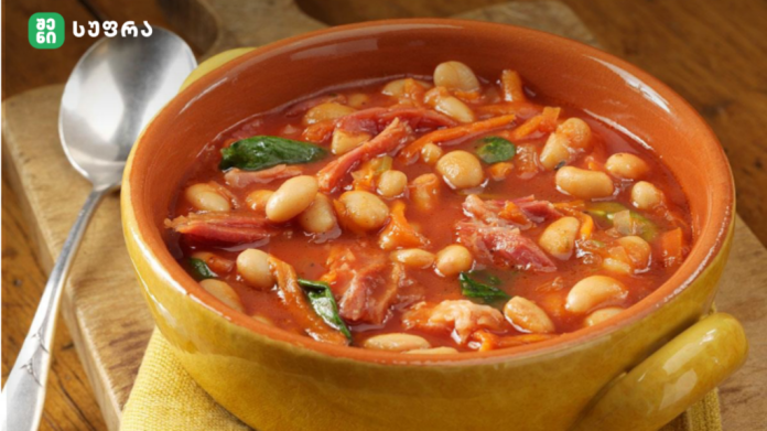 Yellow ceramic bowl holds a hearty white bean and tomato soup with greens on a wooden table, spoon nearby.