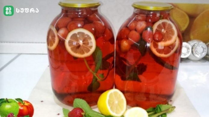 Two large glass jars filled with a red fruit-infused drink with lemon slices and berries, on a white countertop; foreground shows lemons, mint, strawberries, and apples.