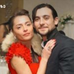 Couple posing together at a formal event; woman in a red dress with a fur stole and the man in a dark suit, background shows a table with flowers and candles.