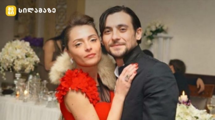 Couple posing together at a formal event; woman in a red dress with a fur stole and the man in a dark suit, background shows a table with flowers and candles.