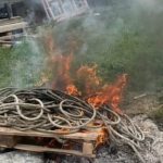 Coiled hoses on a wooden pallet burning in a small outdoor fire with smoke rising nearby debris and grass area on a construction site.