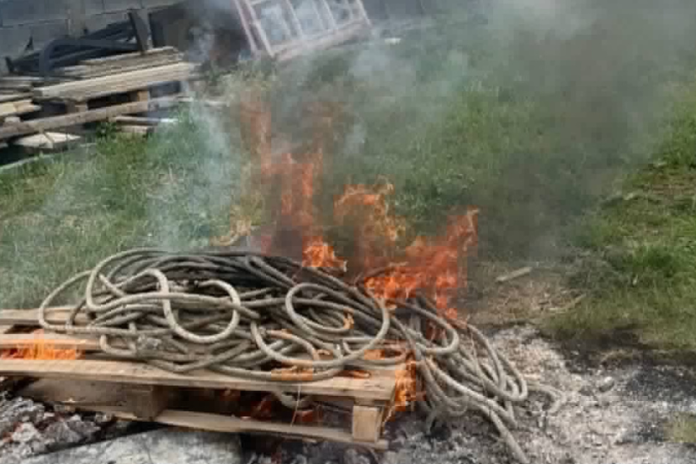 Coiled hoses on a wooden pallet burning in a small outdoor fire with smoke rising nearby debris and grass area on a construction site.