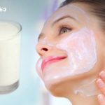 Woman smiles as she applies a white facial mask to her cheek, with a glass of milk visible to the left.