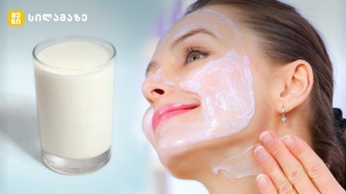 Woman smiles as she applies a white facial mask to her cheek, with a glass of milk visible to the left.
