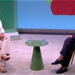 Two people sit in armchairs on a TV talk show set, with a small green table between them and two water glasses on it.
