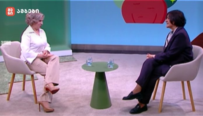 Two people sit in armchairs on a TV talk show set, with a small green table between them and two water glasses on it.