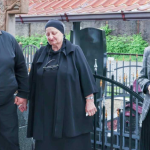 Three adults stand by a grave in a cemetery; a man and an elderly woman in black hold hands, while a woman in a checkered blazer stands nearby holding her hands together.