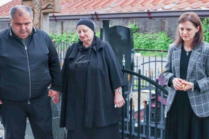 Three adults stand by a grave in a cemetery; a man and an elderly woman in black hold hands, while a woman in a checkered blazer stands nearby holding her hands together.