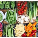 Wide assortment of fresh vegetables laid out on a wooden table, including broccoli, tomatoes, peppers, beans, carrots, and potatoes.